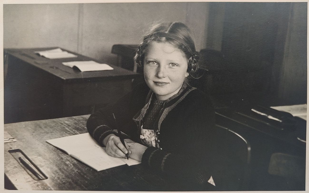 Ein junges Mädchen sitzt an einem alten Schultisch, in der Hand einen Stift, vor sich ein geöffnetes Heft und schaut aufmerksam lächelnd in die Kamera.