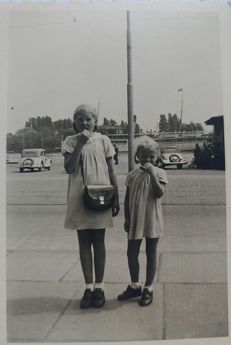 Zwei junge Mädchen in kurzen Sommerkleidchen, nebeneinander stehend, jede mit einer Hand am Mund, auf einem großen, mit Platten belegten Platz, wahrscheinlich ein Bahnhofsvorplatz. Im Hintergrund sieht man zwei Automobile.