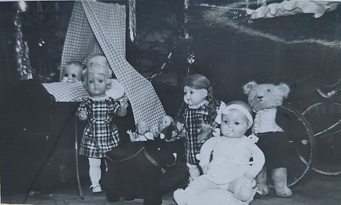 Ein sehr dunkles Foto, auf dem drei größere, sowie einige kleine Puppen und ein Teddybär zu sehen sind. Einige Puppen liegen in einer sehr schlichten Wiege, über der ein karierter Stoff hängt. Puppen und Teddy tragen Kleider, dem Teddy fehlt ein Auge.