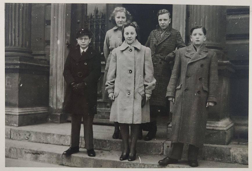 Fünf Jugendliche, zwei Mädchen und drei Jungen in Winterkleidung auf den Stufen vor einer großen Hauseingangstür. Sie schauen sehr ernst in die Kamera.