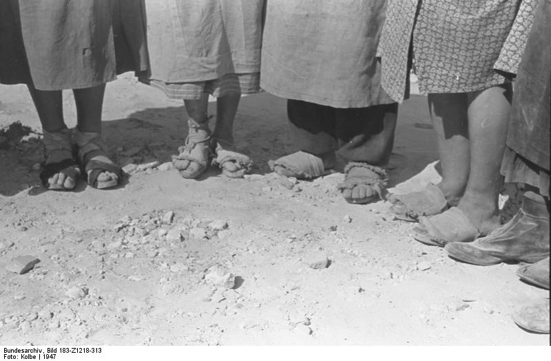 Sechs Frauen stehen in einem Halbkreis, man sieht nur ihre Füße auf dem staubigen Boden und den unteren Rand ihrer Kittelschürzen. Nur zwei von ihnen tragen geschlossene Schuhe, die anderen mehr oder weniger zerschlissene Sandalen oder Latschen. Nicht unbedingt zueinander passende.