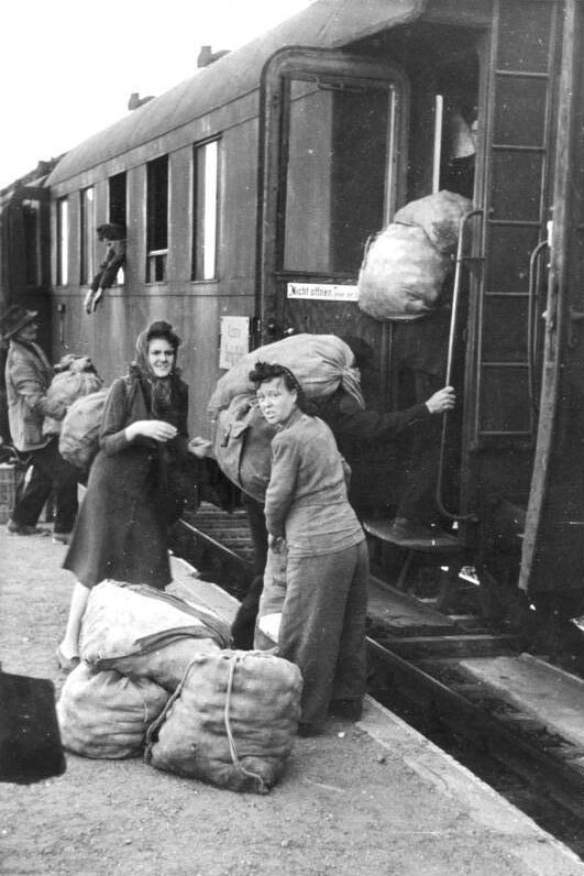 Zwei Frauen stehen neben einigen Säcken mit Kartoffeln vor der offenen Tür eines Zuges, in den Menschen durch Fenster und Türen Säcke mit Kartoffeln hieven.