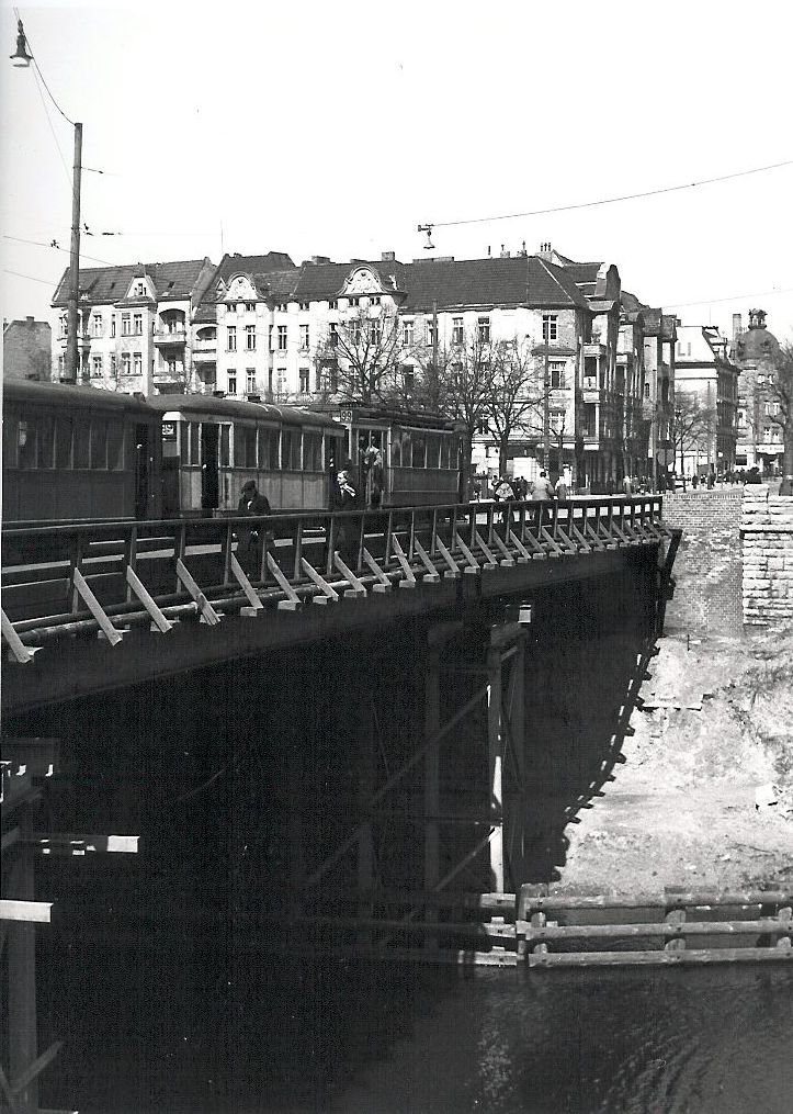 Eine Brücke auf einer Balkenkonstruktion über einen Kanal, darauf eine Straßenbahn mit drei Wagen. Das Ufer fällt über gut zehn Meter sehr steil zum Wasser hin ab. Im Hintergrund sieht man ein großbürgerliches, vierstöckiges Eckgebäude.