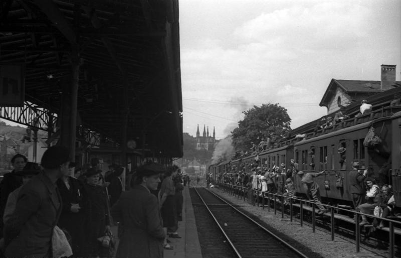 In einem Bahnhof steht rechts ein völlig überfüllter Zug, die Menschen sitzen auf dem Dach und auf den Puffern. Links warten Menschen an einem Bahnsteig.